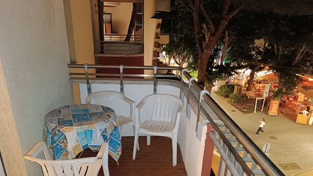 Een balkon met een tafel, stoelen, een leuning en uitzicht op een straat met winkels en mensen.