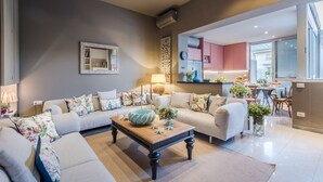 Un salon moderne avec un ensemble de canapés, une table basse et un coin cuisine visible à travers une porte ouverte.