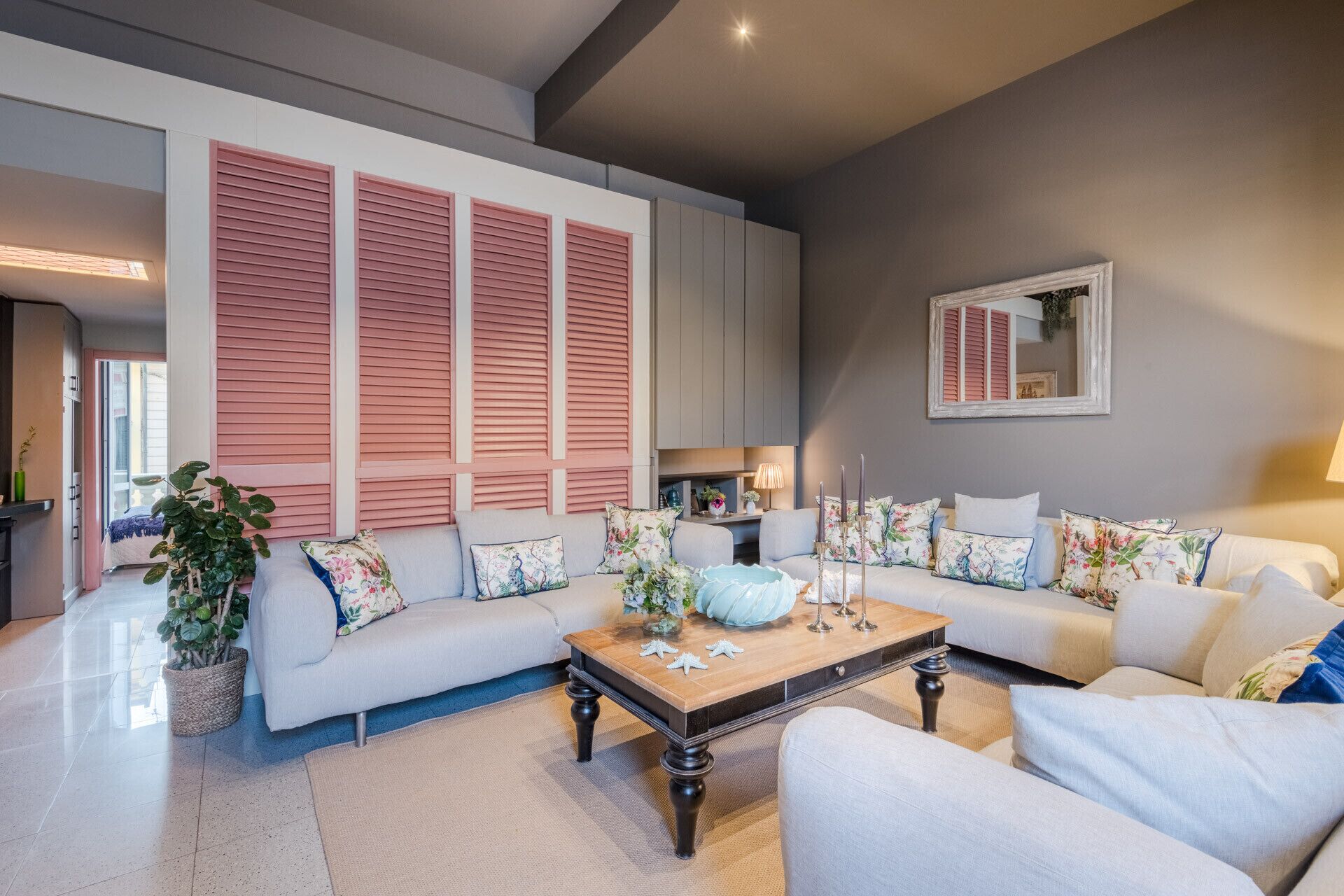 Uma sala de estar ampla com um grande sofá branco, uma mesa de centro de madeira e uma janela grande com persianas cor-de-rosa.