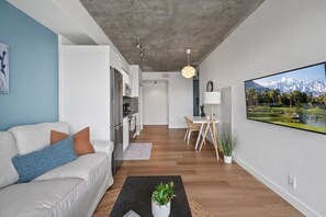 Un salon moderne avec un canapé, un coin repas comprenant une table et des chaises, une cuisine et un grand téléviseur diffusant un paysage de montagne.