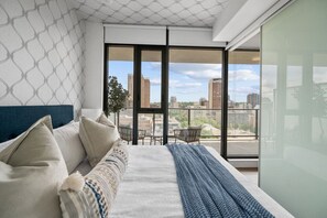 Une chambre moderne dotée d’un grand lit, offrant une vue sur la ville grâce à de grandes fenêtres du sol au plafond, et d’un balcon aménagé avec du mobilier d’extérieur.