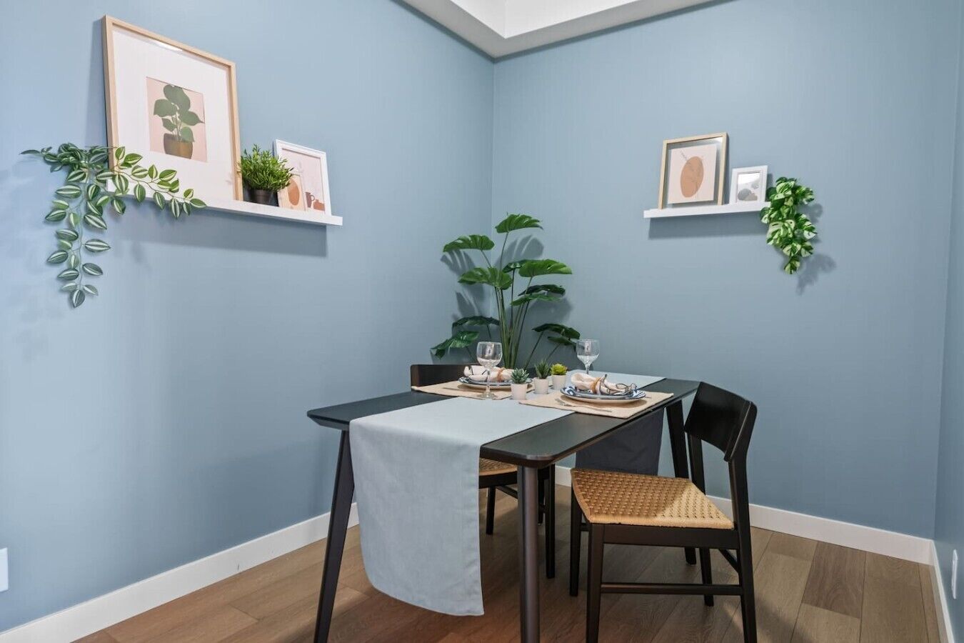 Een eetgedeelte met een tafel voor twee personen, omringd door stoelen, met een blauwe wand en planken met ingelijste foto's en planten.