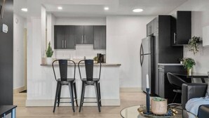 Una cocina moderna con armarios oscuros, una isla central y dos sillas. Hay un comedor con mesa y sillas, y una mesita redonda con un adorno.