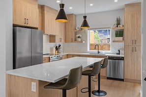 A modern kitchen with wooden cabinets, a stainless steel refrigerator, a marble island with bar stools, and pendant lighting.