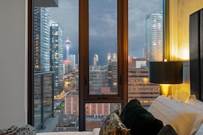 Une chambre dotée d’une grande fenêtre donnant sur la ville, d’un lit avec des oreillers et d’une lampe fixée au mur.