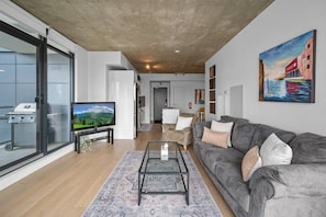 A modern living room with a flat-screen TV, a grey sofa, a coffee table, and a large window.