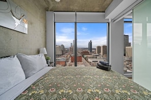 A bedroom with a large bed, a view of a cityscape through floor-to-ceiling windows, and a concrete ceiling.