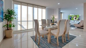 A modern living room with a glass dining table, comfortable chairs, a large window, and a flat-screen TV.