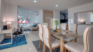 A modern living room with a glass dining table, beige chairs, a blue rug, a TV, and a kitchen in the background.