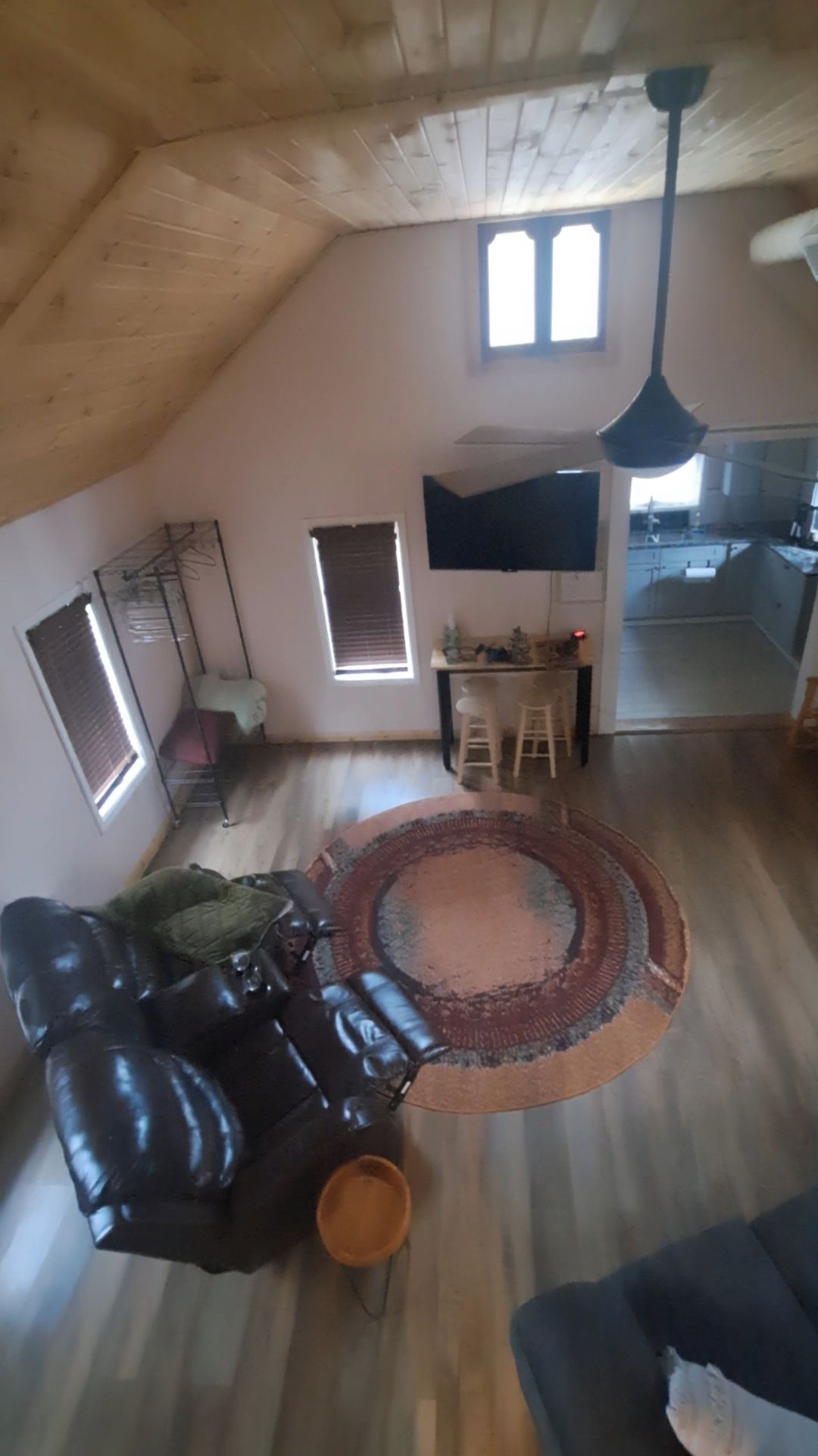 Une chambre mansardée avec un plafond en bois, un canapé en cuir noir, un tapis rond et un coin repas avec des tabourets.