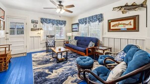 A living room with a blue sofa, a wooden coffee table, and wicker chairs. There is a ceiling fan, a fish decoration on the wall, and a framed fish picture.