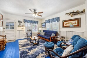 A living room with a blue sofa, a wooden coffee table, and wicker chairs. There is a ceiling fan, a fish decoration on the wall, and a framed fish picture. - Historic * Walkable * Downtown * Boat Parking * Fire Pit (Cedar Key)