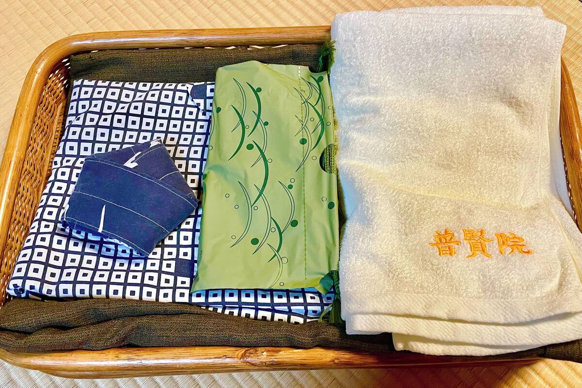 Een netjes opgeruimde verzameling items in een houten mand, waaronder een gestreept kussen, een groene doek, een witte handdoek met tekst, en een donkerblauwe muts.