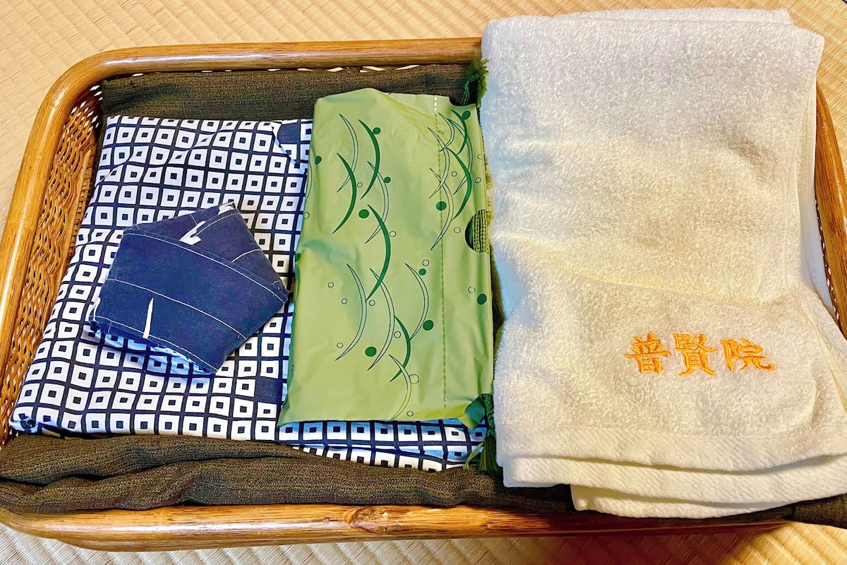 Un ensemble d’objets soigneusement disposés dans un panier en bois, comprenant un coussin à motifs, un tissu vert, une serviette blanche avec du texte et une casquette bleu foncé.