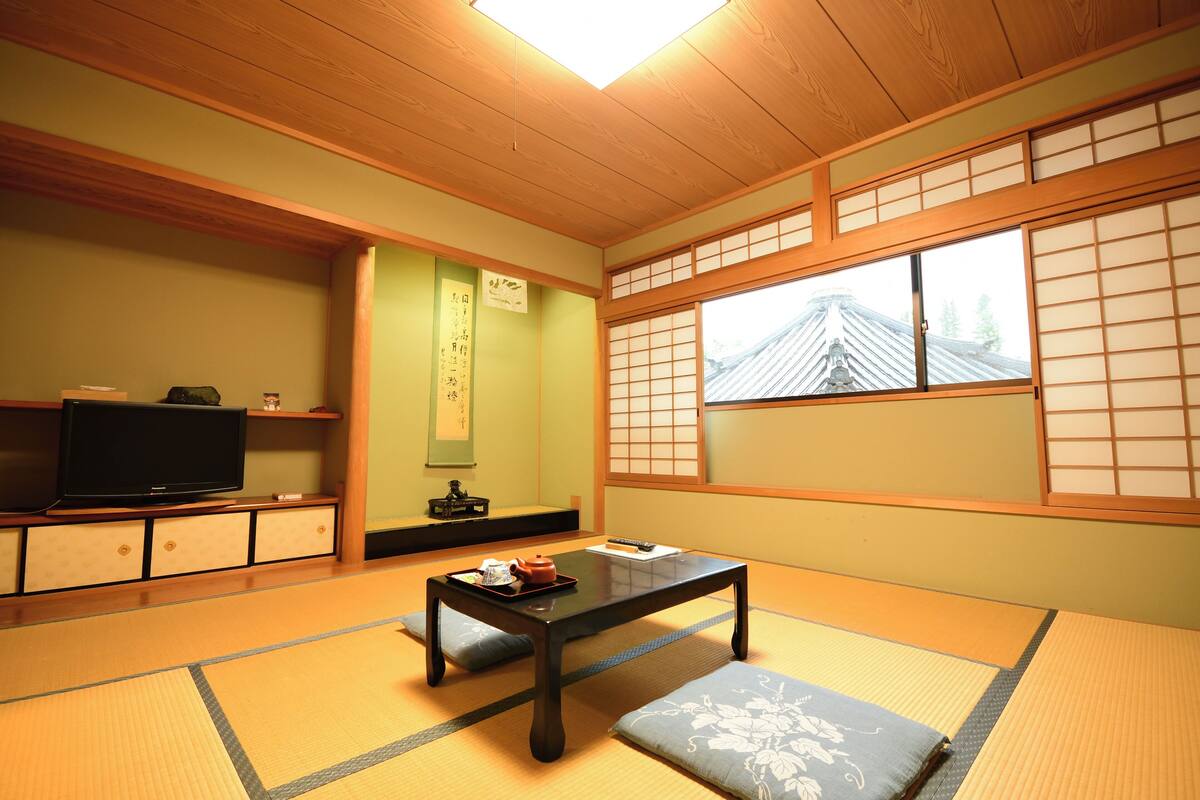 Een traditionele Japanse kamer met tatami vloerbedekking, een lage tafel, kussens, een televisie en schuifdeuren.