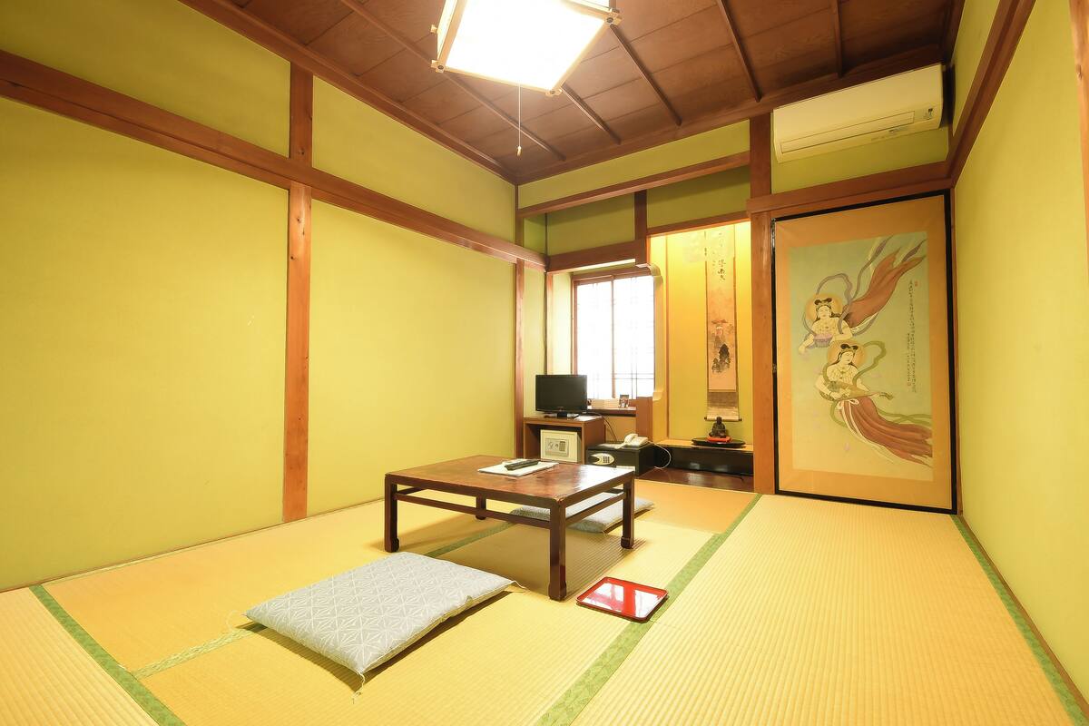Een kamer met tatami matten, een lage houten tafel, een kussen, een rode dienblad, een televisie en een lijst met een foto aan de muur.