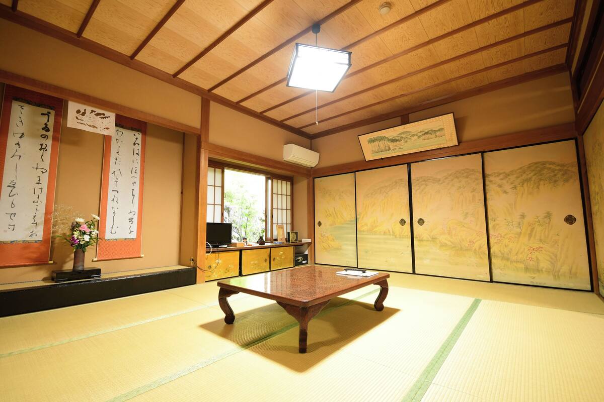 Een kamer met tatami matten, een houten tafel, schuifdeuren met kunstwerk, een plafondventilator en een raam met natuurlijk licht.