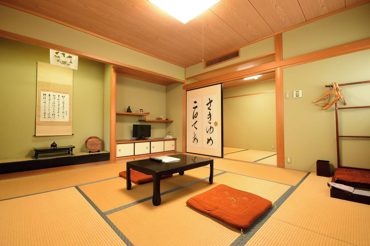 Een traditionele Japanse kamer met tatami vloerbedekking, een lage tafel, kussens, een televisietoestel en kalligrafie op de muren.