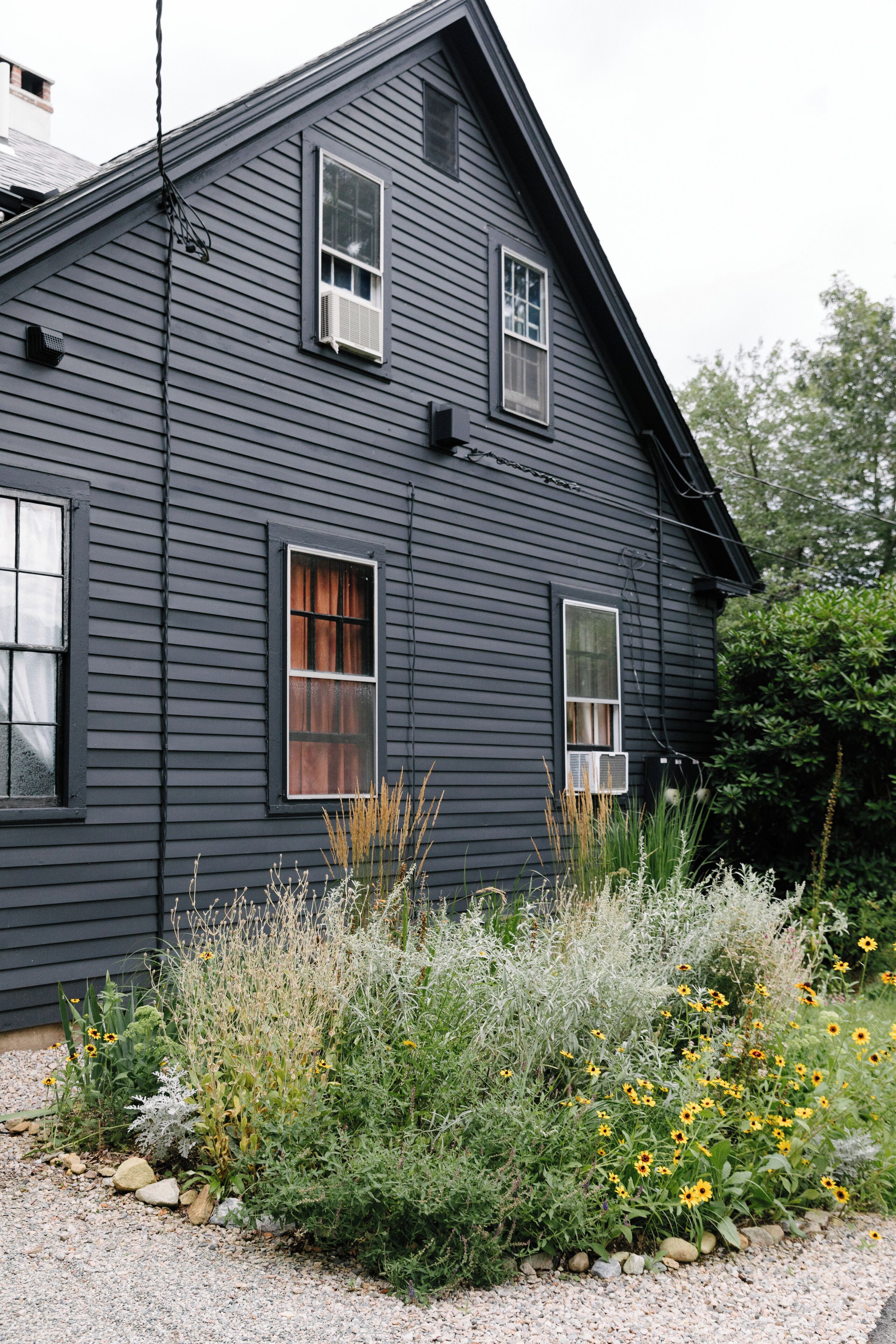 A two-story house with a dark exterior, two visible windows with curtains, and a gravel driveway with various plants and flowers.
