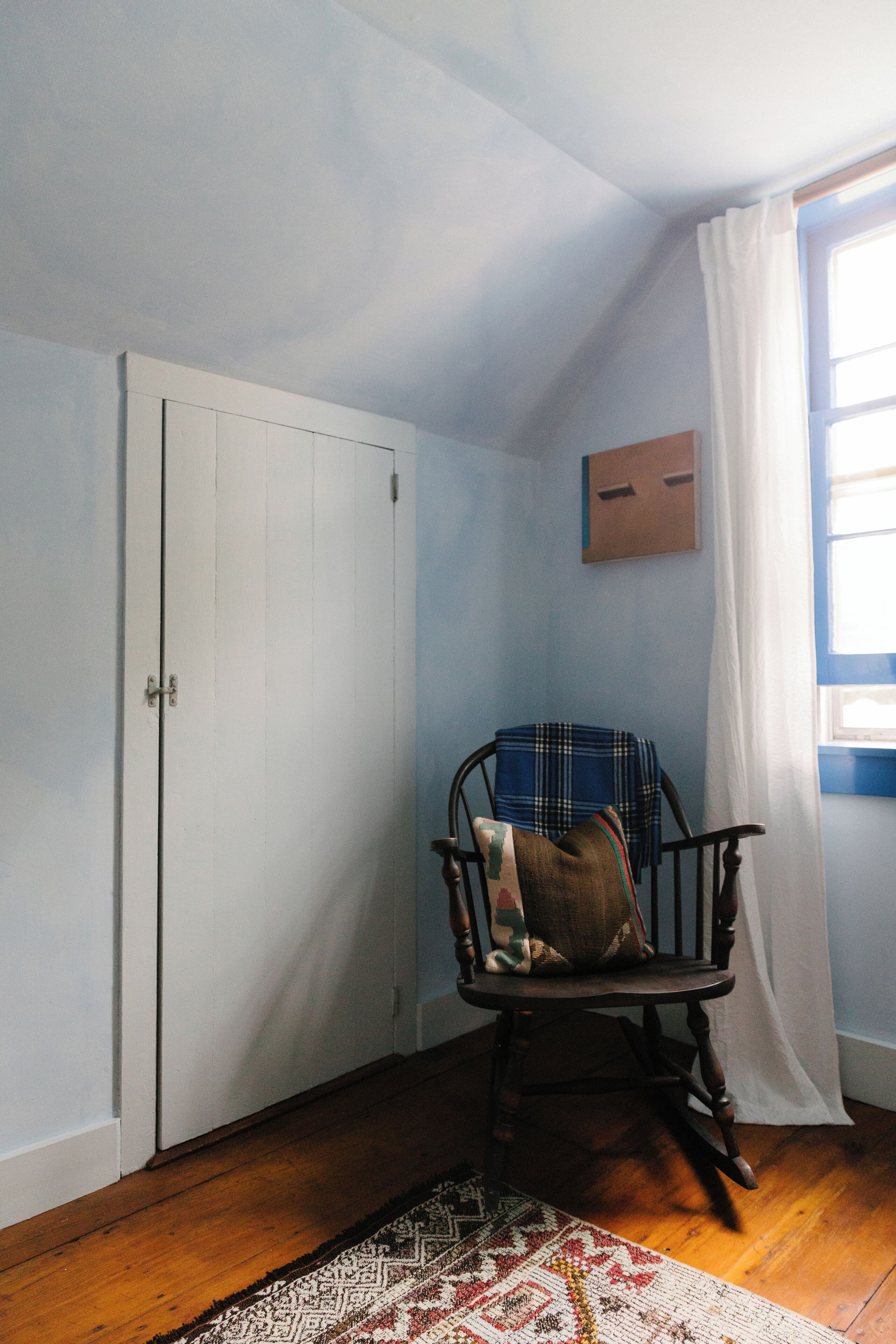 A room with a wooden floor, a white door, a chair with a plaid blanket, and a window with white curtains.
