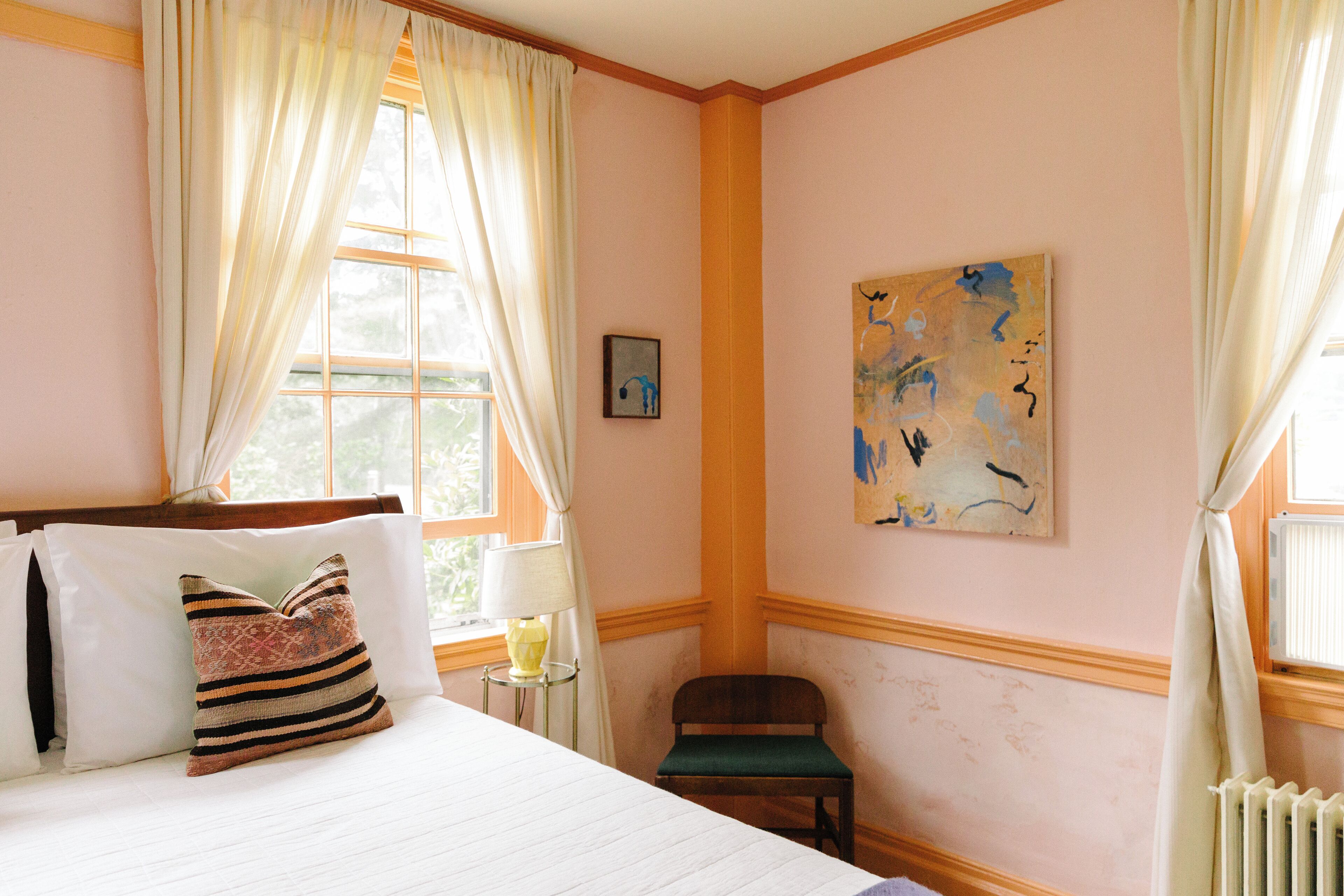 Ein Schlafzimmer mit Bett, Kissen, einem Sessel, einem kleinen Tisch mit Lampe und einem Bild an der Wand.