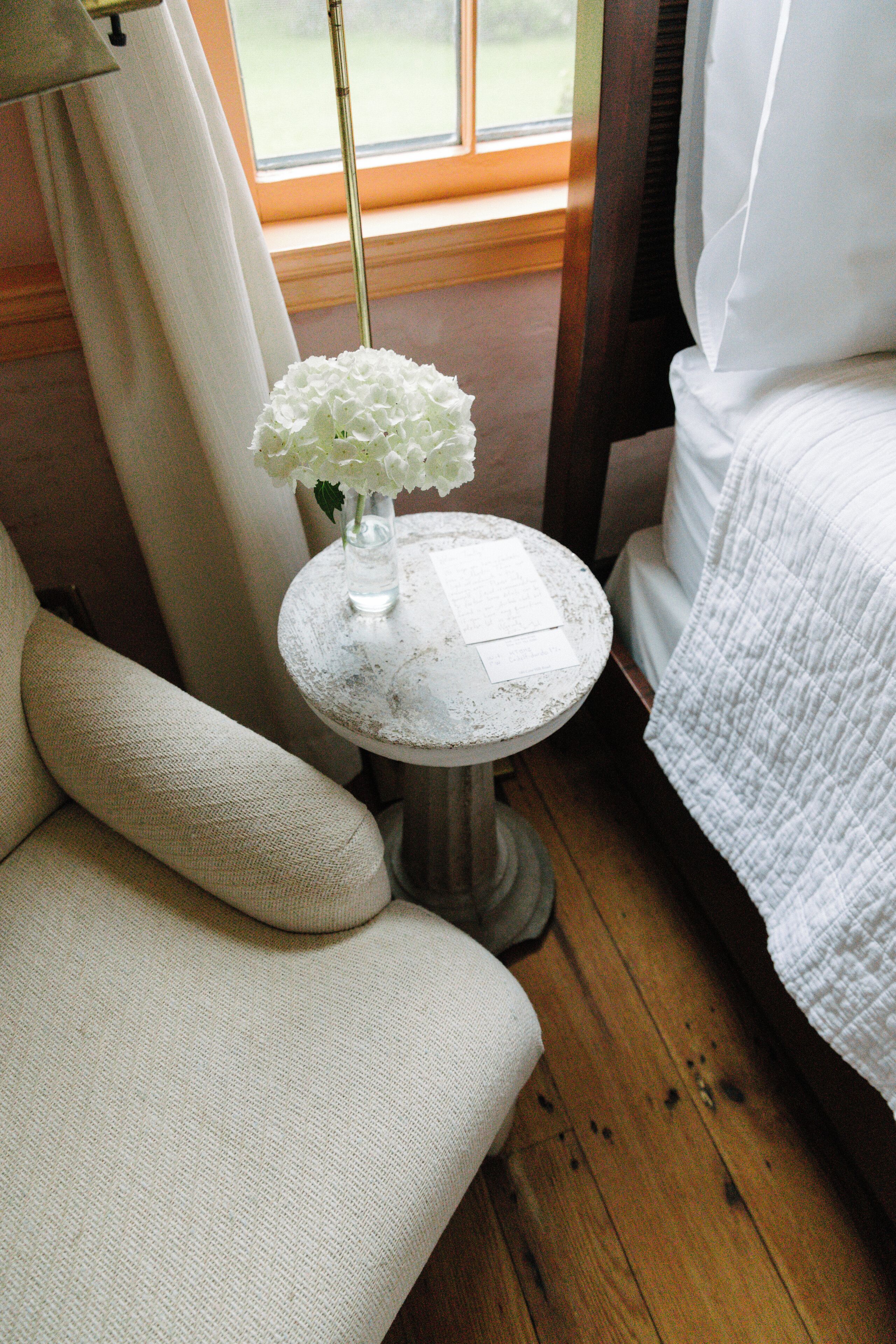 A room with a white upholstered sofa, a small round table with a vase of white flowers, and a bed with white bedding.