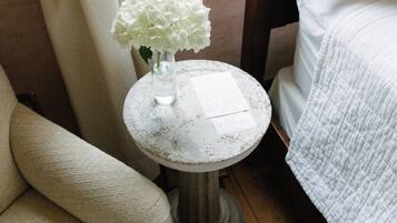 A room with a white upholstered sofa, a small round table with a vase of white flowers, and a bed with white bedding.