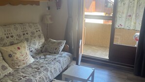 A living room with a sofa, two small tables, a rug, wooden cabinets with heart-shaped cutouts, a lamp, and a view through a sliding glass door. - Studio Montagne 20 m (Saint-Léger-les-Mélèzes)