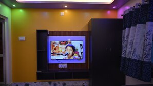 Een woonkamer met een flatscreen televisie aan een wand, een donkere entertainmentkast eronder en vloertegels met een patroon.