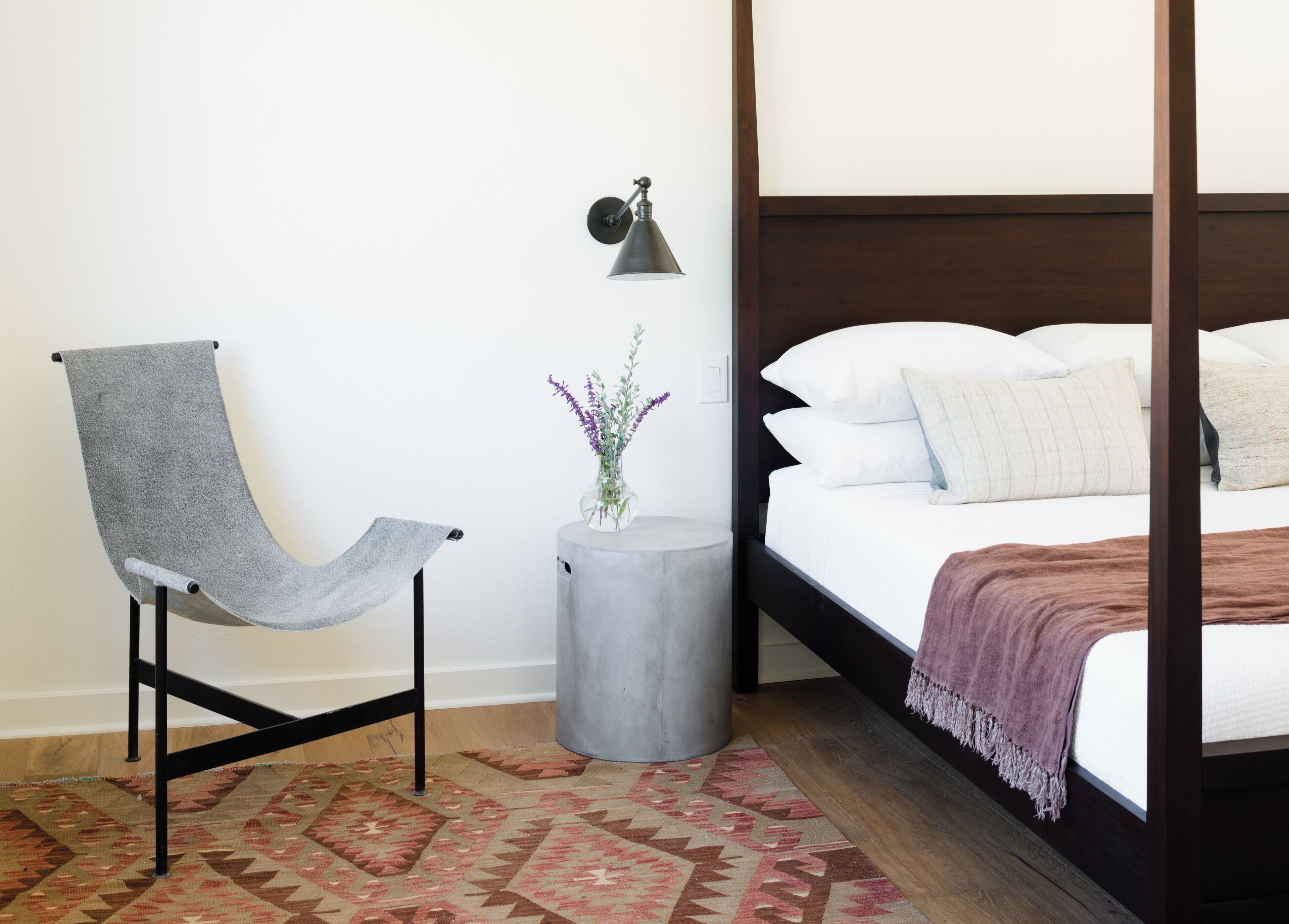 A bedroom with a four-poster bed, a grey upholstered chair, a small round table with a vase of flowers, and a patterned rug.