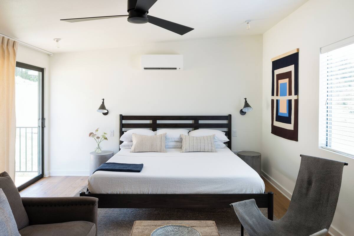 Une chambre à coucher comprenant un lit, un ventilateur de plafond, un climatiseur, une table de chevet, une lampe et une fenêtre avec des stores.