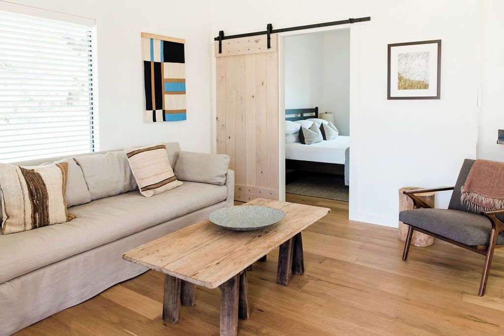 Uma sala de estar moderna com sofá, mesa de centro de madeira, uma cadeira e uma cama ao fundo.