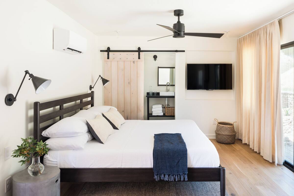 Une chambre moderne avec un lit, des lampes de chevet, un ventilateur de plafond, une télévision et une grande fenêtre avec des rideaux.