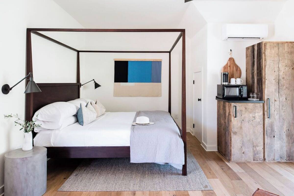 Un lit à baldaquin avec une structure en bois foncé, un matelas blanc et un plaid gris. Une petite table de chevet avec un vase de plantes vertes et une lampe se trouve à côté du lit. Une armoire en bois à l’aspect rustique est placée sur le côté droit.