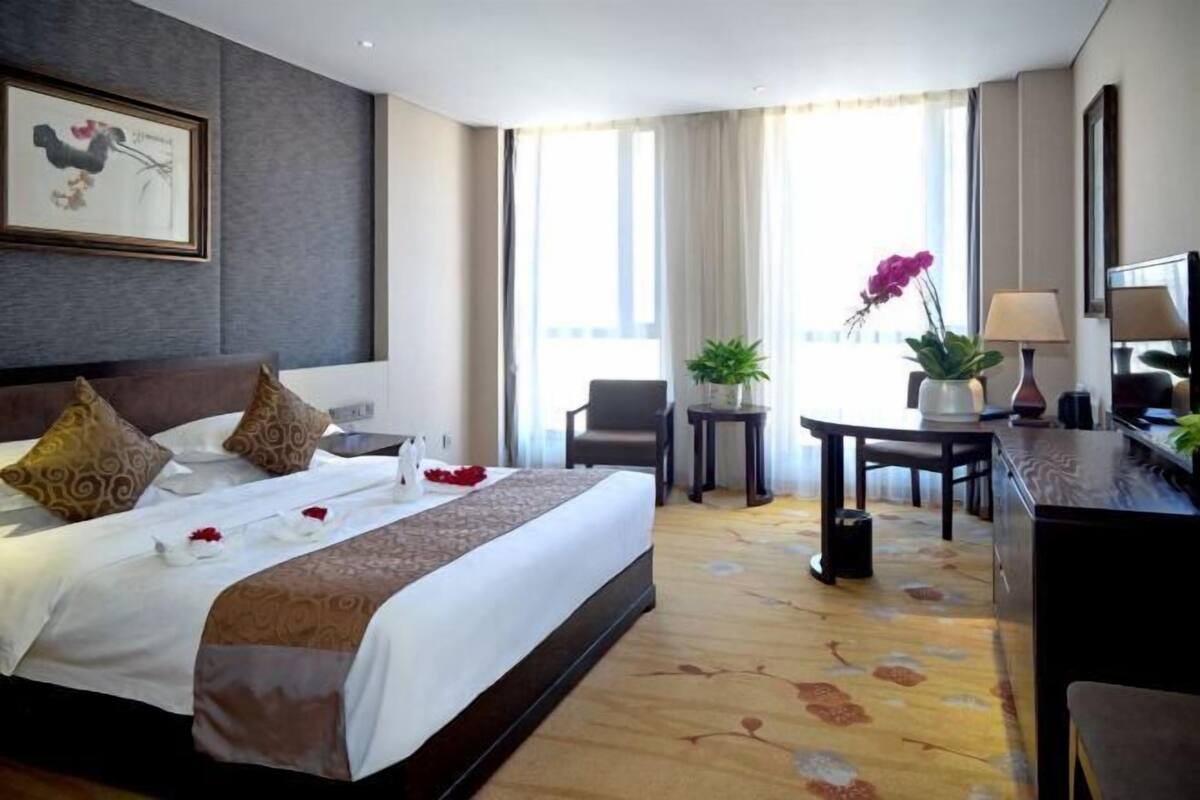 A hotel room with a large bed, a desk, a chair, a small table, and a potted plant.
