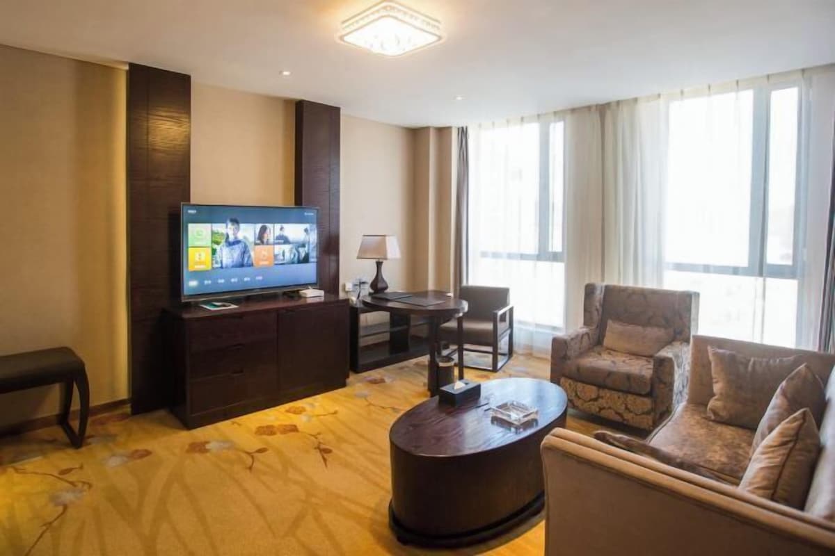 A hotel room with a flat-screen TV, a sofa, a coffee table, a lamp, and a large window with curtains.