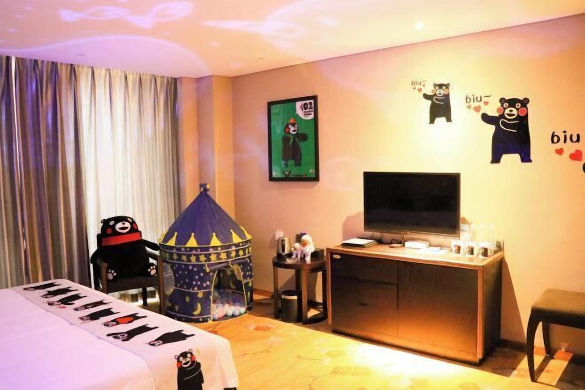 A hotel room with a bed, a TV, a desk, a chair, a small play tent, and wall decorations including cartoon bears and hearts.