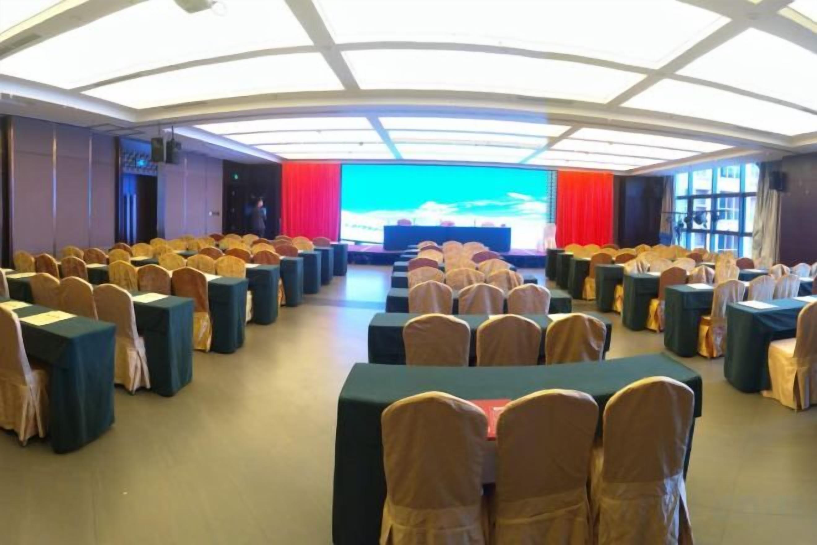 A conference room with rows of tables and chairs, a stage with a large screen, and a high ceiling with skylights.