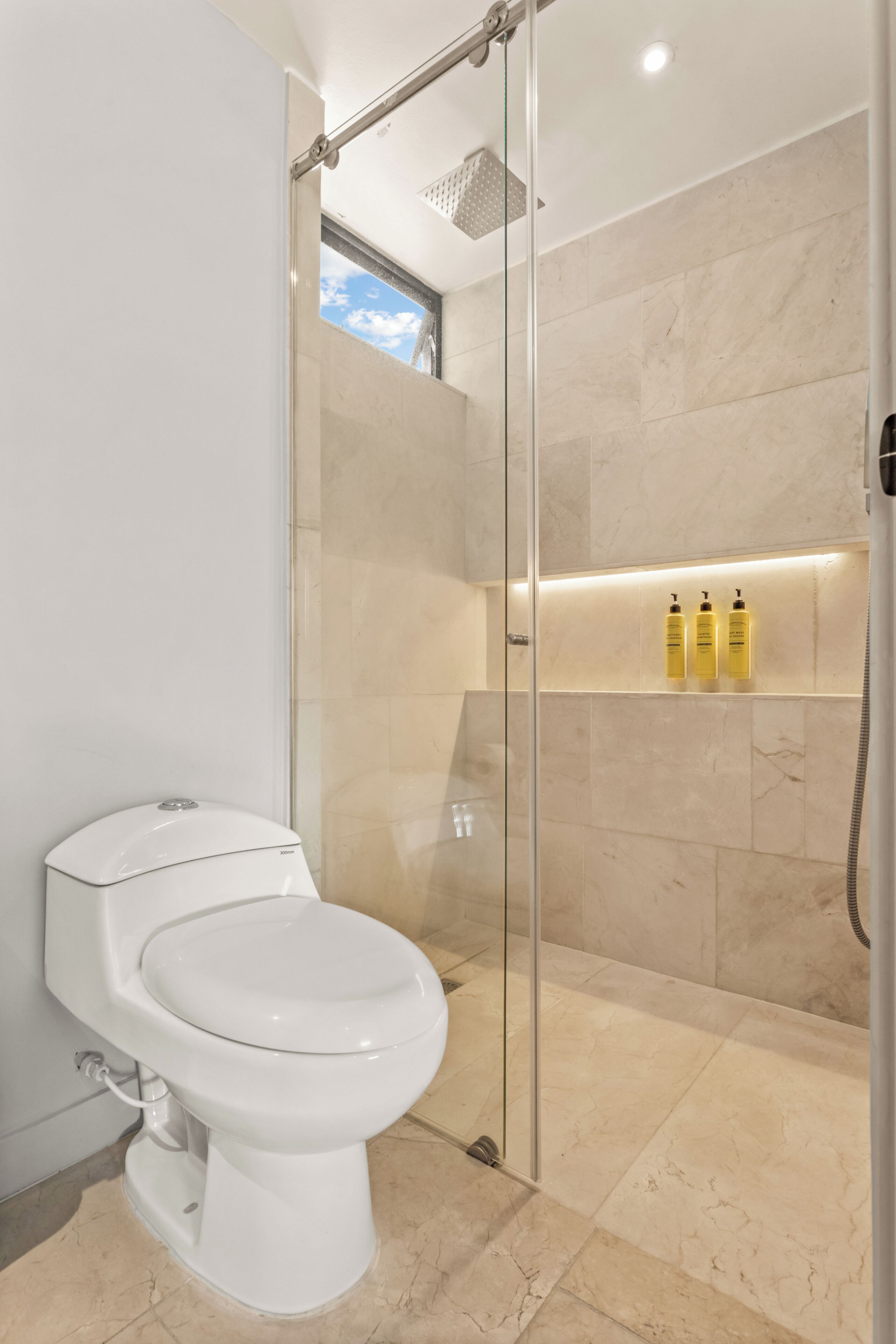 Un baño moderno con ducha de vidrio, inodoro y estante con artículos de aseo.