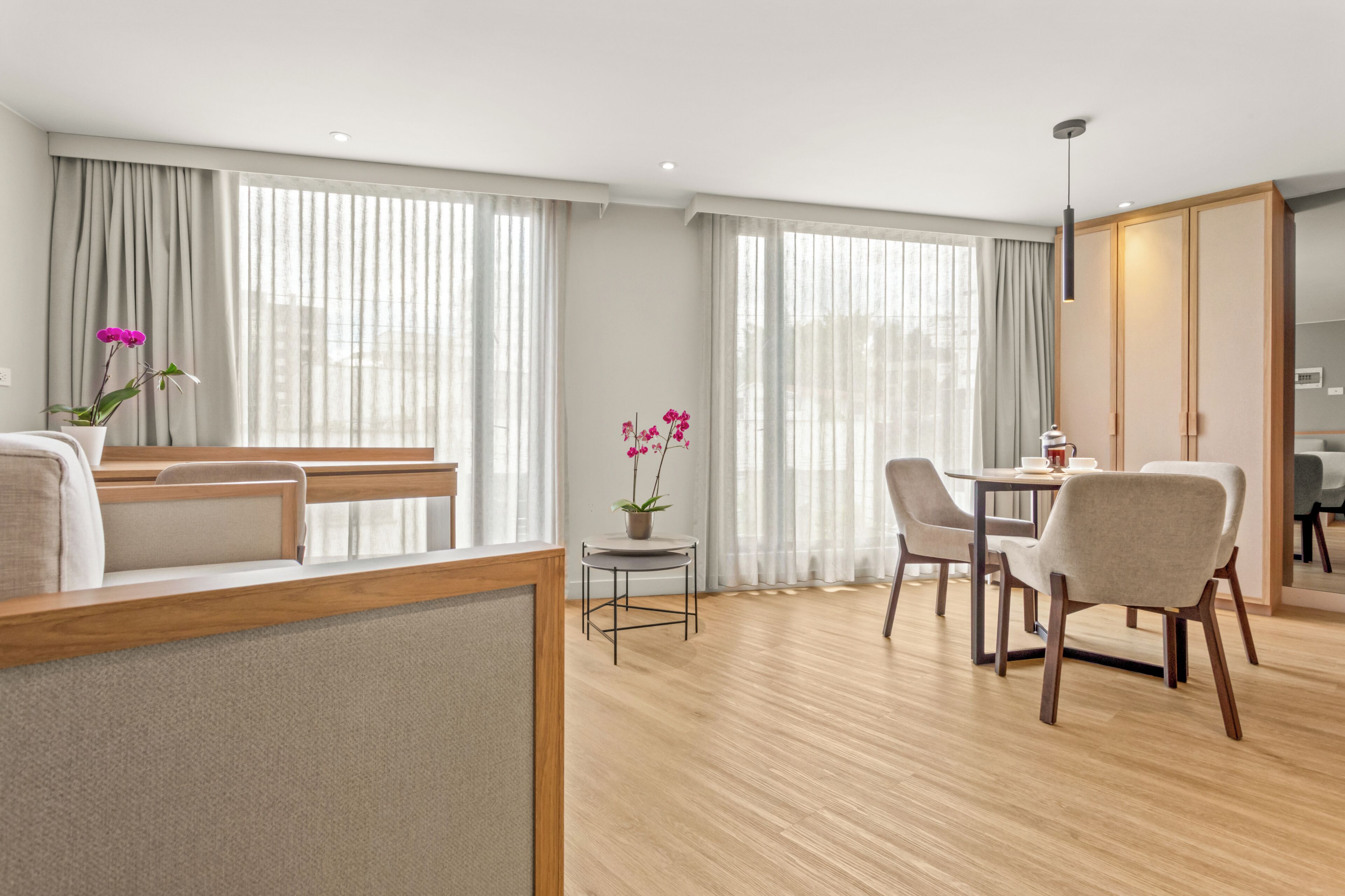 Quarto de hotel moderno com piso de madeira, área de refeições com mesa e cadeiras, e uma grande janela com cortinas.