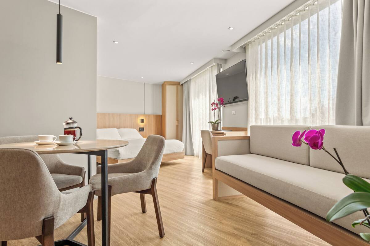 A modern hotel room with a dining area, a sofa, a TV, and a large window with curtains.