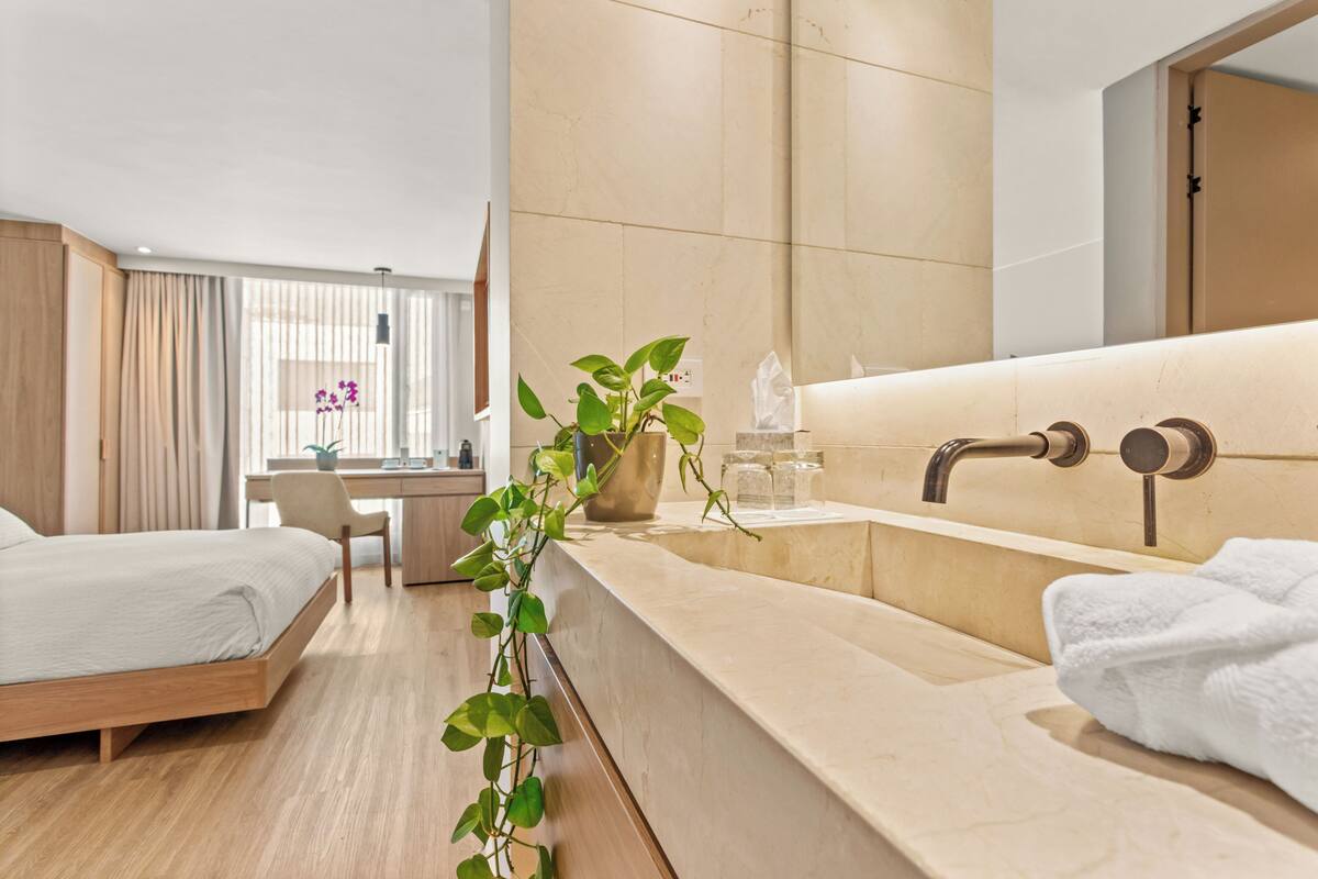 A modern hotel room with a bed, a desk, a bathroom with a large mirror, and a plant.