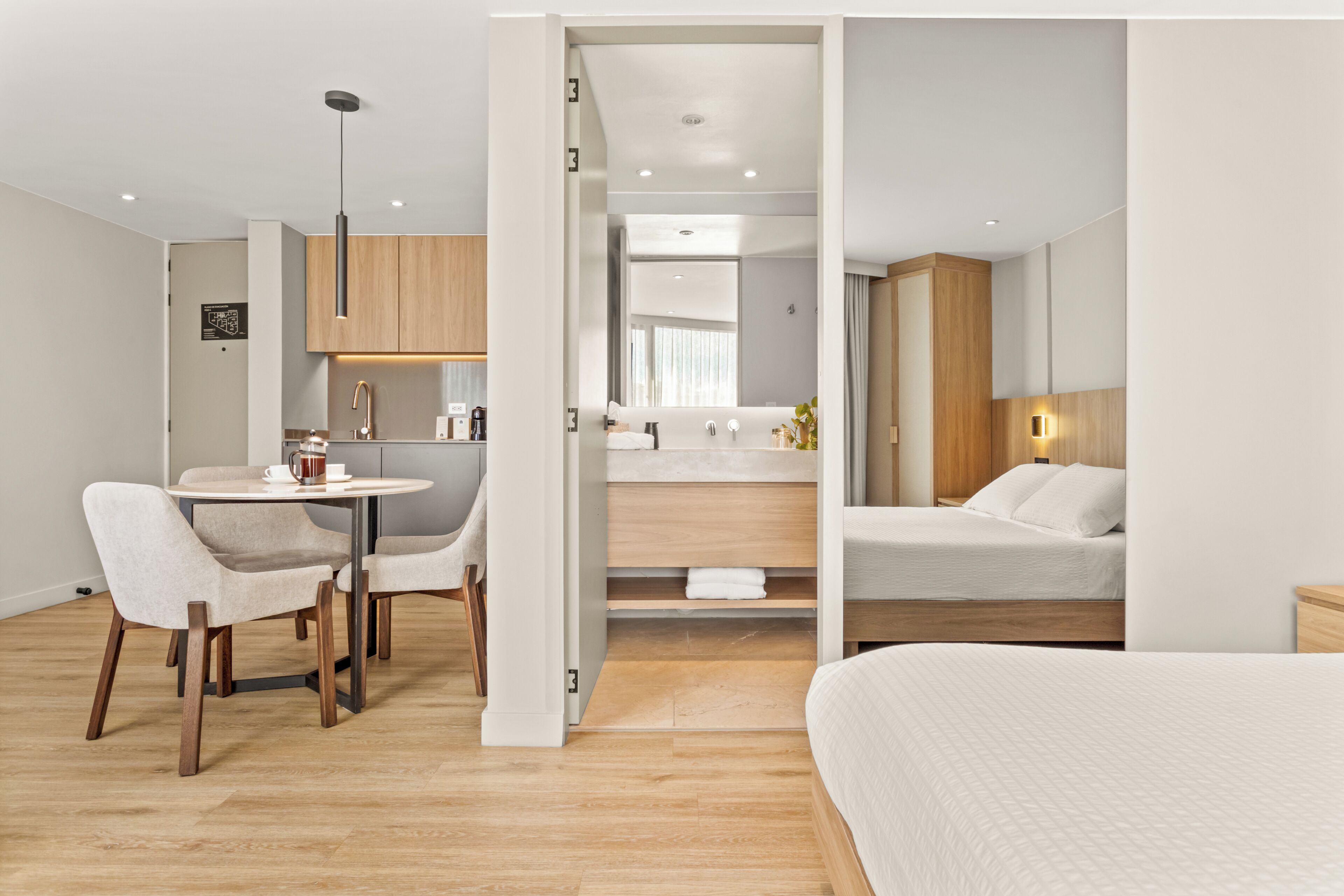 A modern hotel room with a bed, a dining area with chairs and a table, a kitchenette, and a bathroom with a mirror and sink.