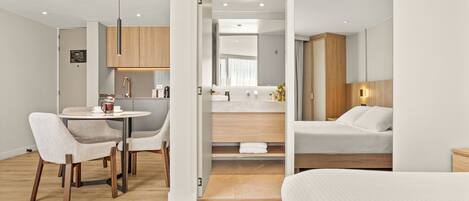 A modern hotel room with a bed, a dining area with chairs and a table, a kitchenette, and a bathroom with a mirror and sink.