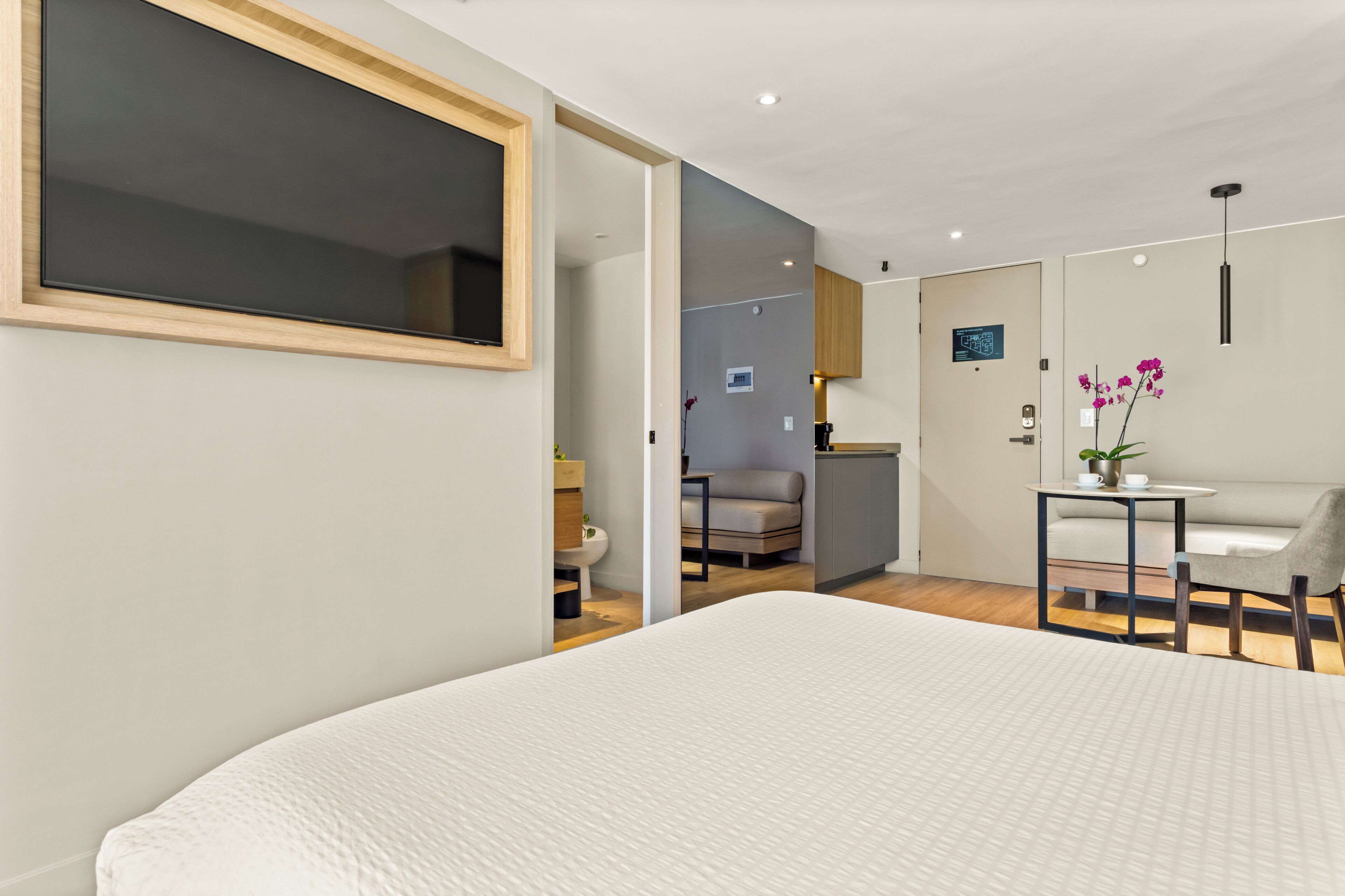Una habitación de hotel moderna con una cama grande, televisión, una kitchenette pequeña y una zona de estar con una mesa y una silla.