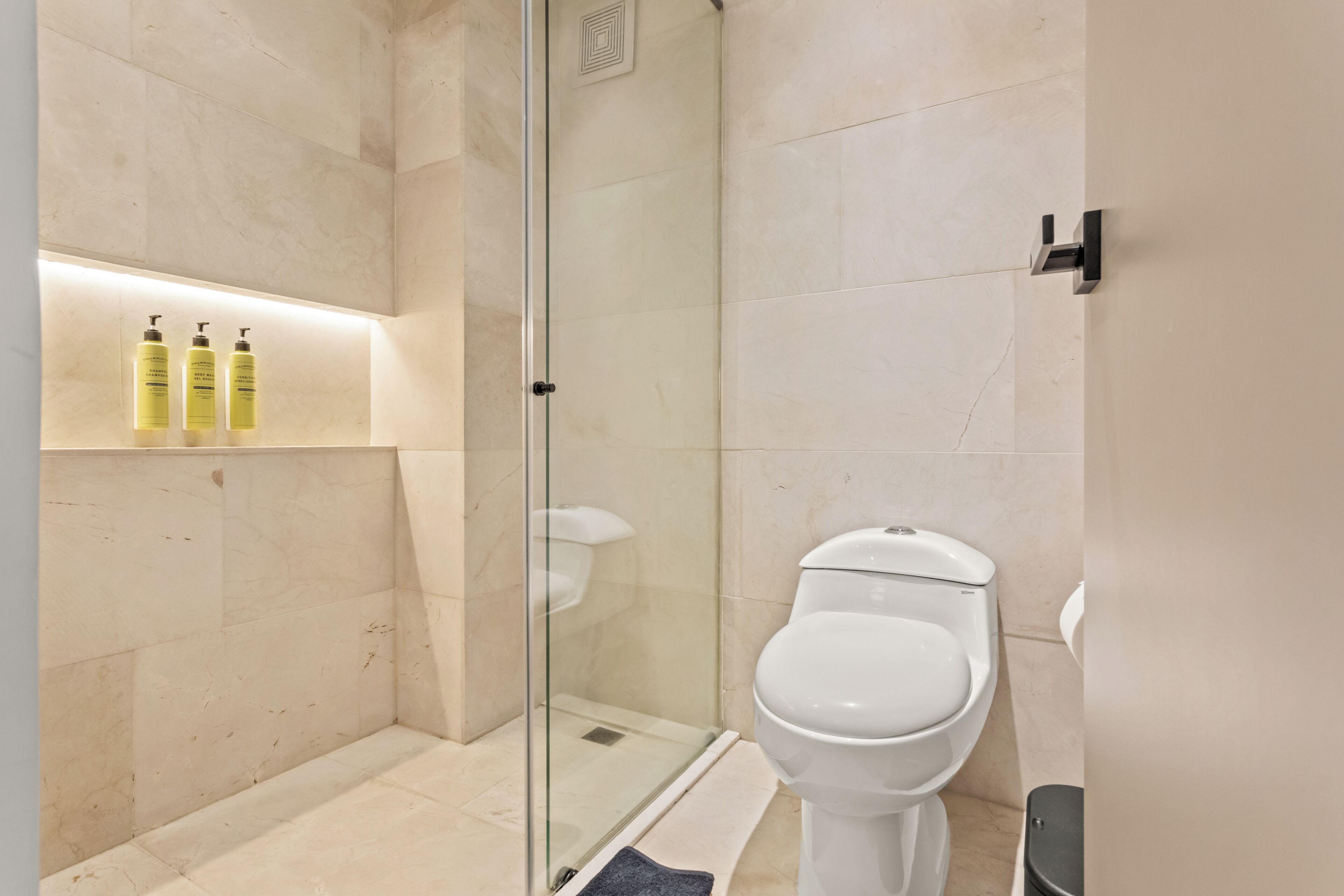 Un baño moderno con ducha de vidrio, inodoro blanco y estante con artículos de aseo.