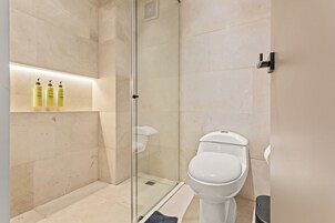Un baño moderno con ducha de vidrio, inodoro blanco y estante con artículos de aseo.
