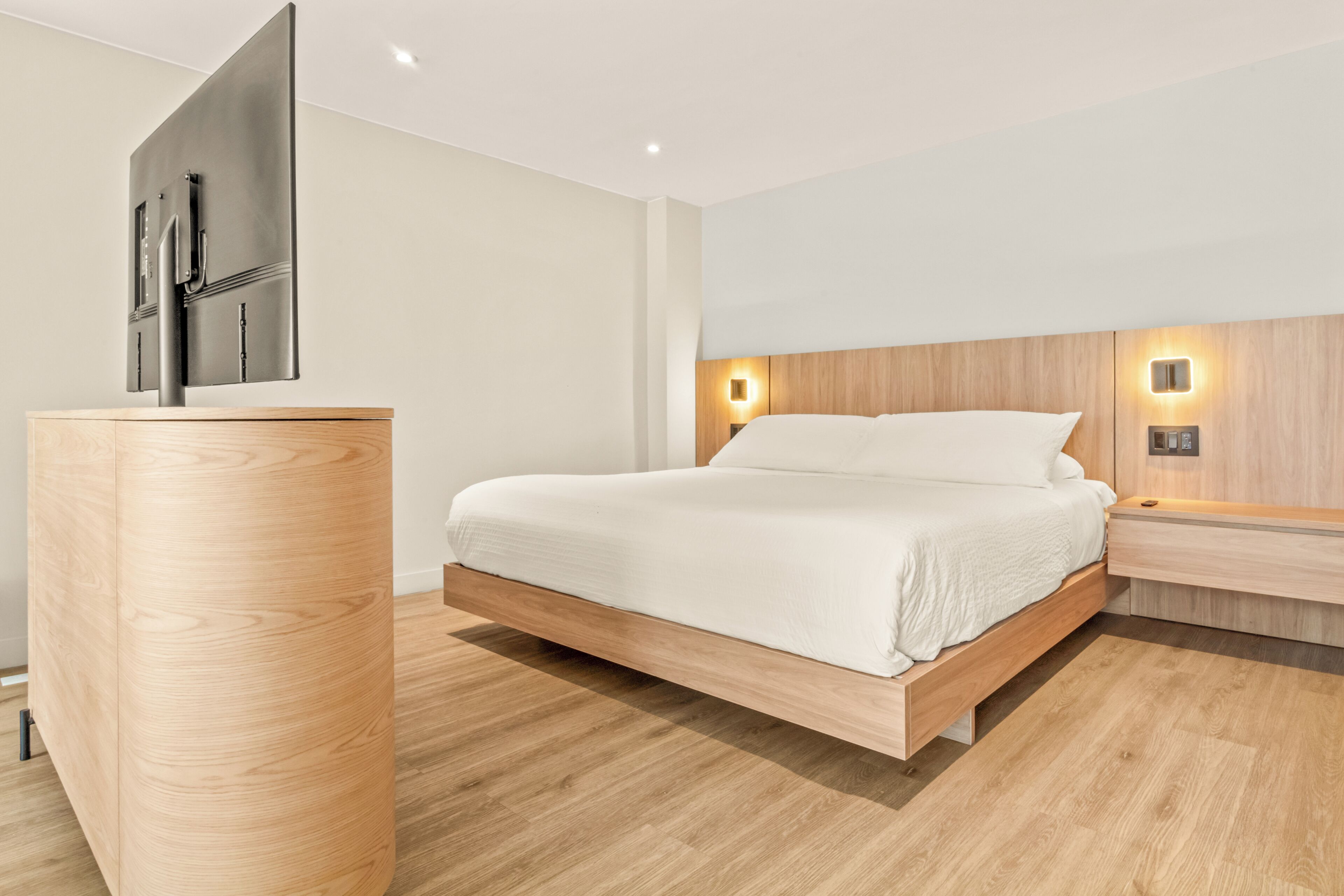 A modern bedroom with a minimalist design, featuring a bed with a wooden frame, a wooden nightstand, a flat-screen TV mounted on the wall, and a wall-mounted light fixture.