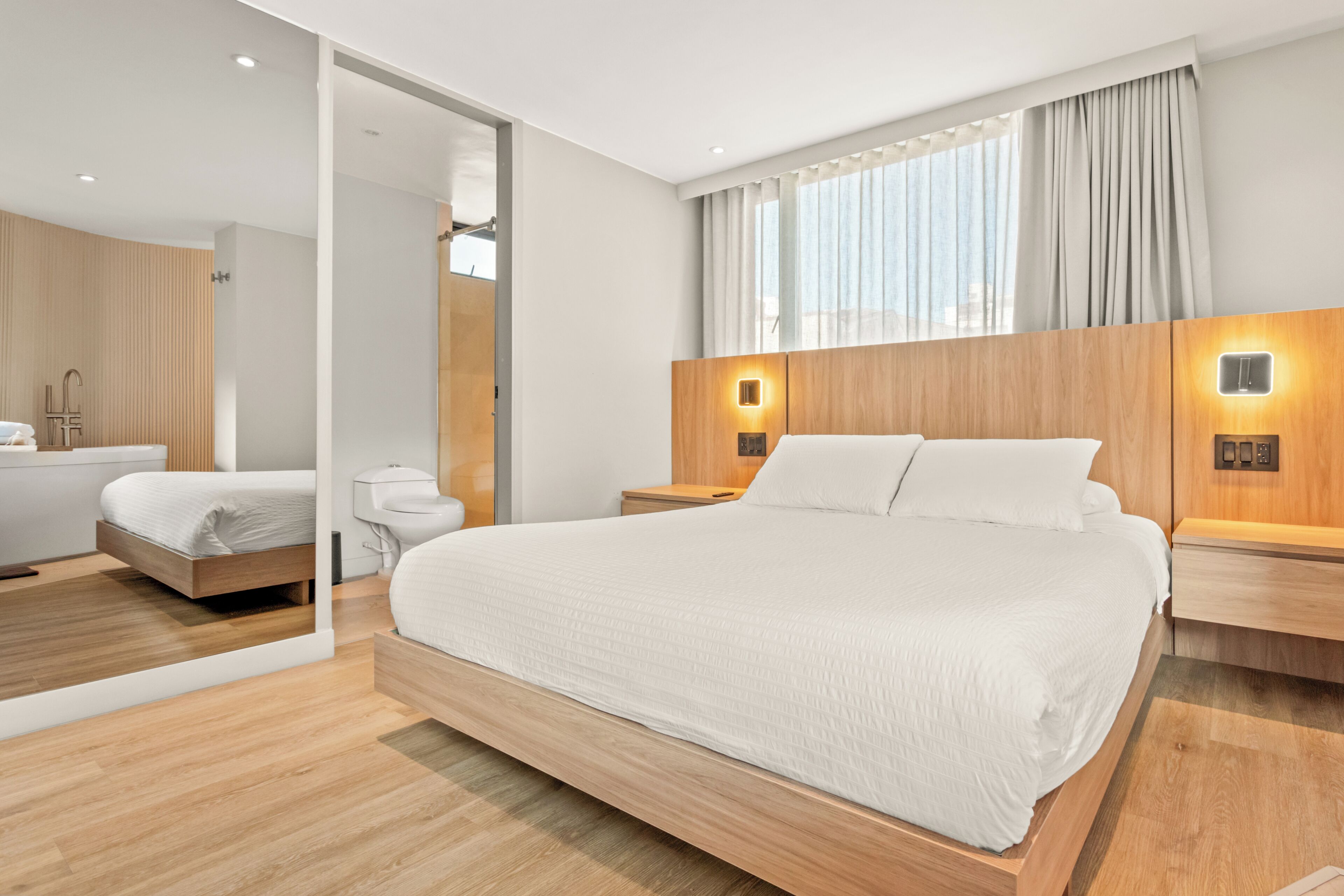 Ein modernes Hotelzimmer mit einem großen Bett, einem Holzkopfteil, Nachttischen, einem Spiegel und einem Badezimmer mit Toilette und Waschbecken.
