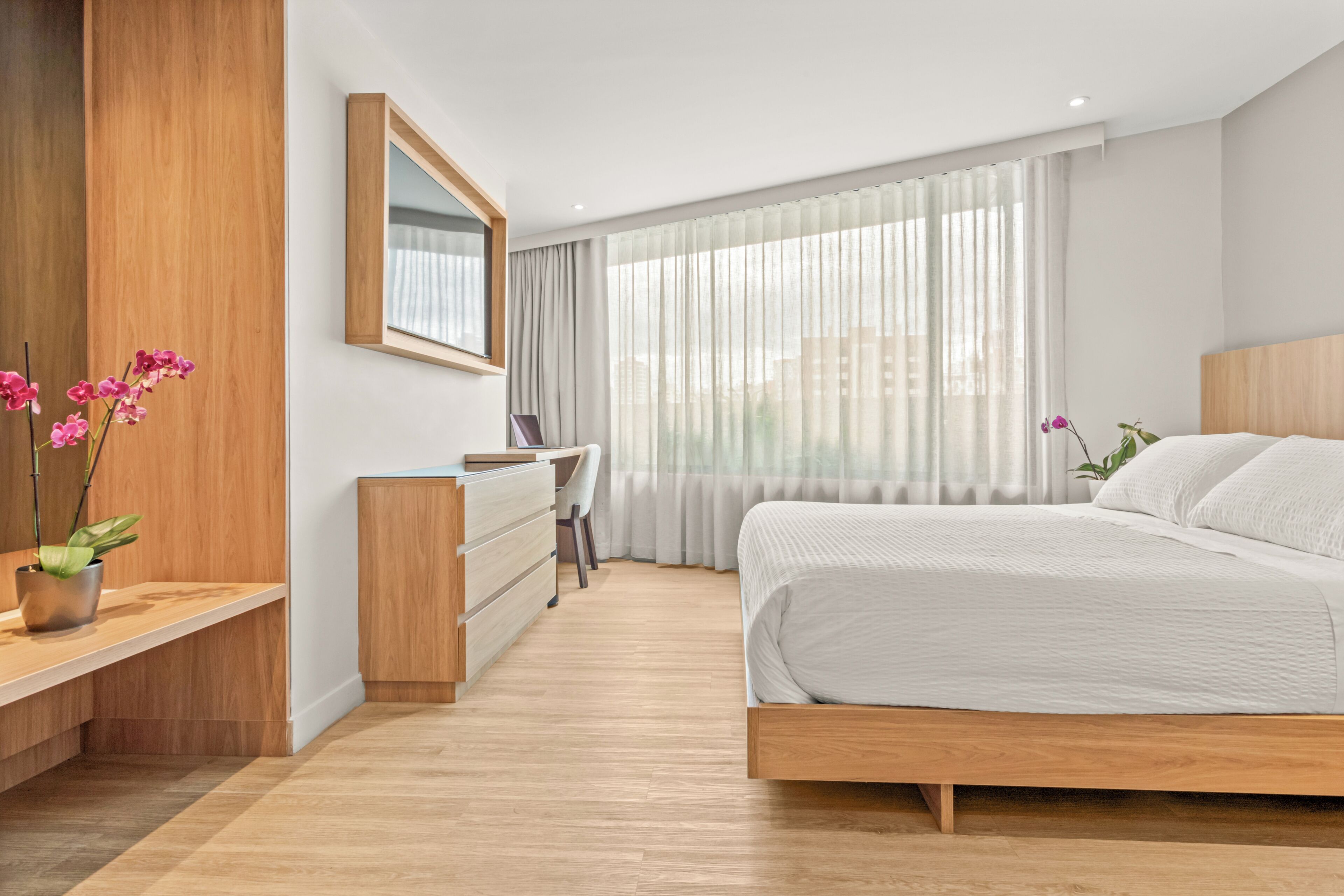 A modern bedroom with a wooden bed, a desk, and a chair, and a large window with sheer curtains.